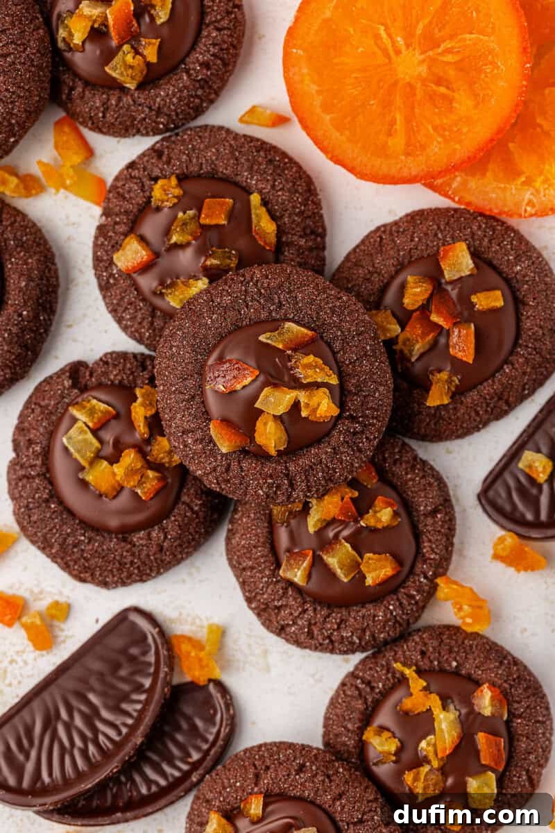A pile of chocolate thumbprint cookies topped with chocolate and small pieces of candied orange, surrounded by chocolate orange slices and fresh orange slices on a light surface.