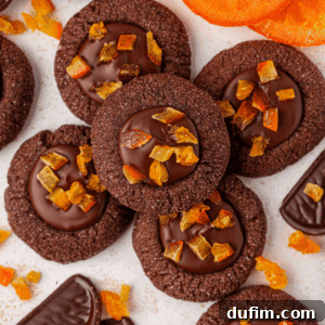 Chocolate thumbprint cookies filled with glossy chocolate and topped with small pieces of candied orange, arranged closely together with scattered orange slices and chocolate pieces nearby.