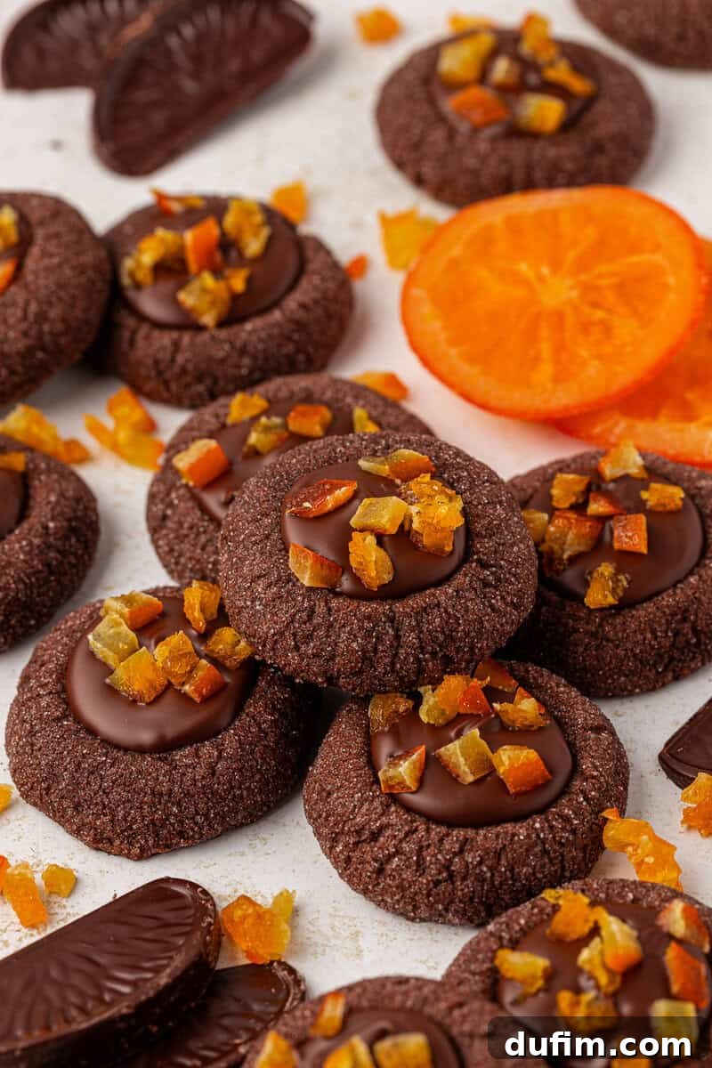 Chocolate thumbprint cookies topped with chocolate and orange peel pieces are arranged on a surface, surrounded by chocolate segments and slices of fresh orange.