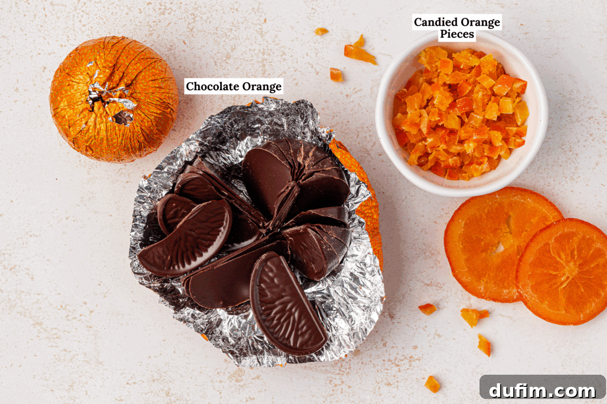 A foil-wrapped chocolate orange is partially unwrapped, showing chocolate orange slices. Nearby are candied orange pieces in a bowl, an unwrapped chocolate orange, and two slices of dried orange.