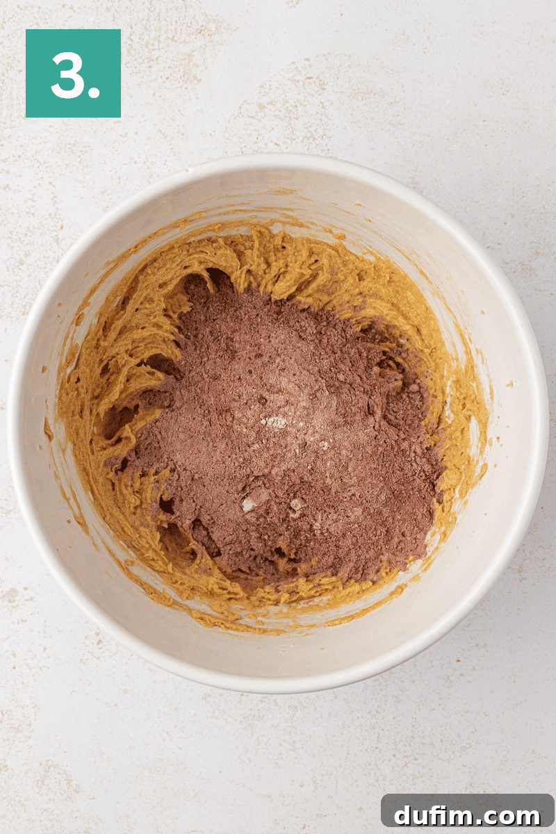 A white mixing bowl with a creamy, light brown batter partially mixed with cocoa powder, sitting on a light surface. A green box with the number 3 is in the top left corner.