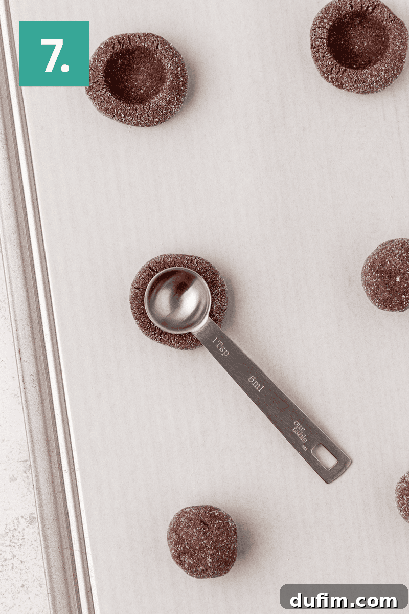 A baking tray holds perfectly portioned chocolate dough balls, each delicately coated in granulated sugar. A metal teaspoon is actively pressing a neat indentation into the center of one dough ball, creating the thumbprint shape. The number 7 in a green box indicates the step.