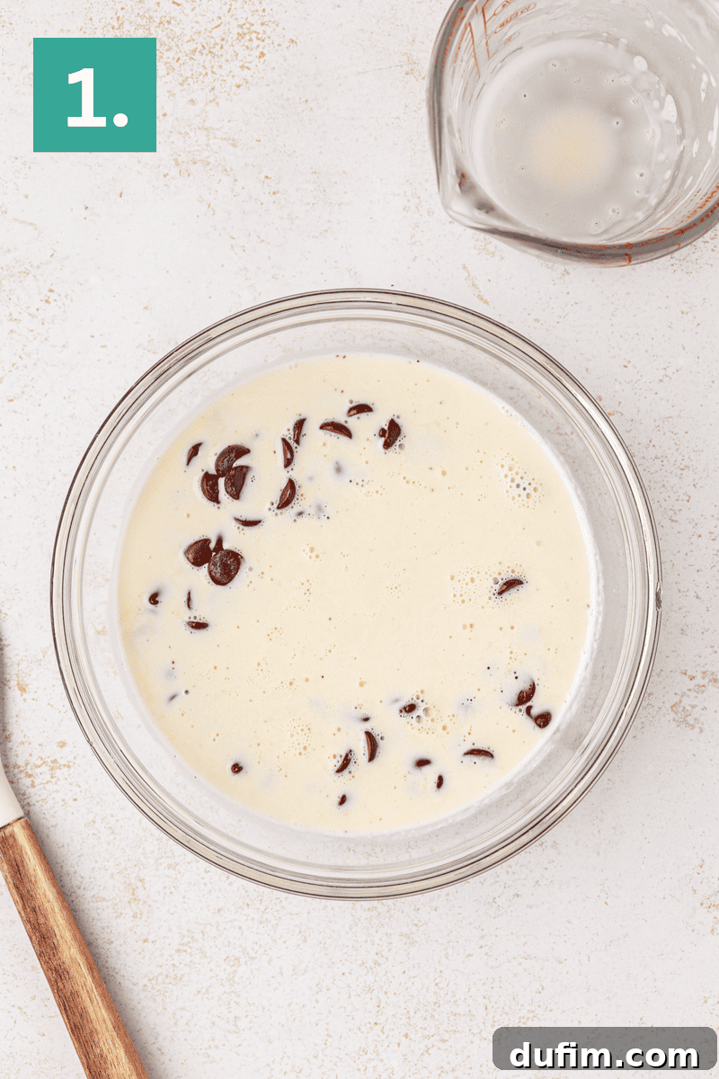 A glass bowl holds semi-sweet chocolate chips, alongside a microwave-safe measuring cup with heavy whipping cream and a wooden-handled spatula, set on a light countertop. A green box with the number 1 indicates the initial step for the filling.