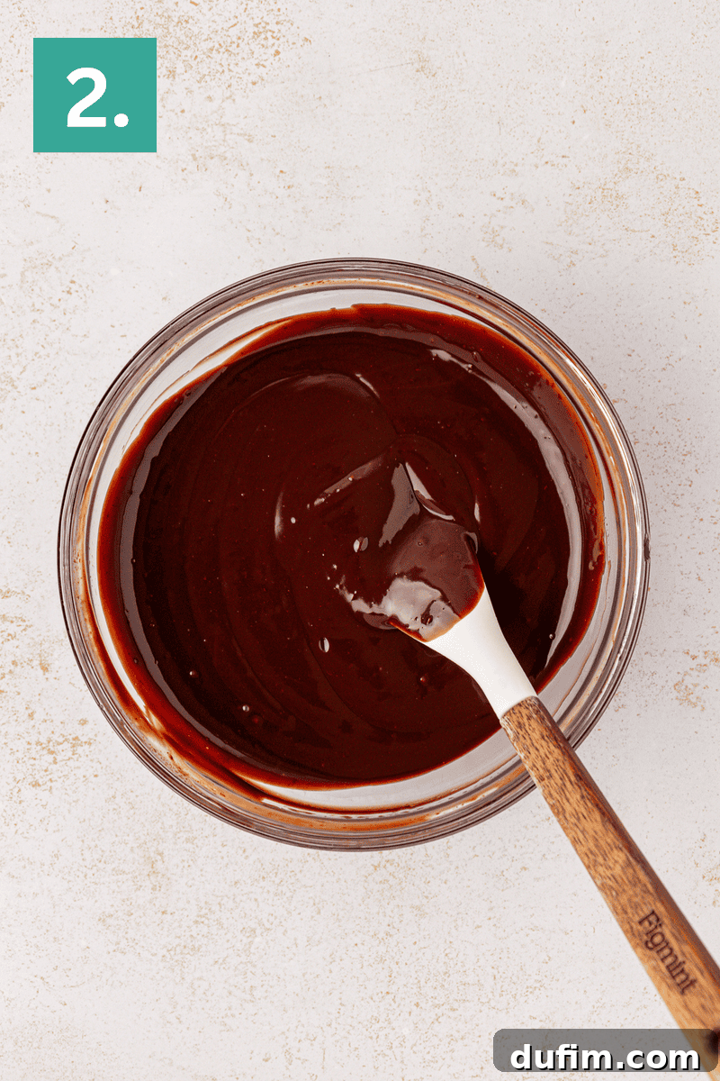 A clear glass bowl is filled with a rich, smooth, and glossy melted chocolate ganache, with a wooden-handled spatula resting inside. The view is from above on a light surface. A green box with the number 2 denotes the subsequent step.