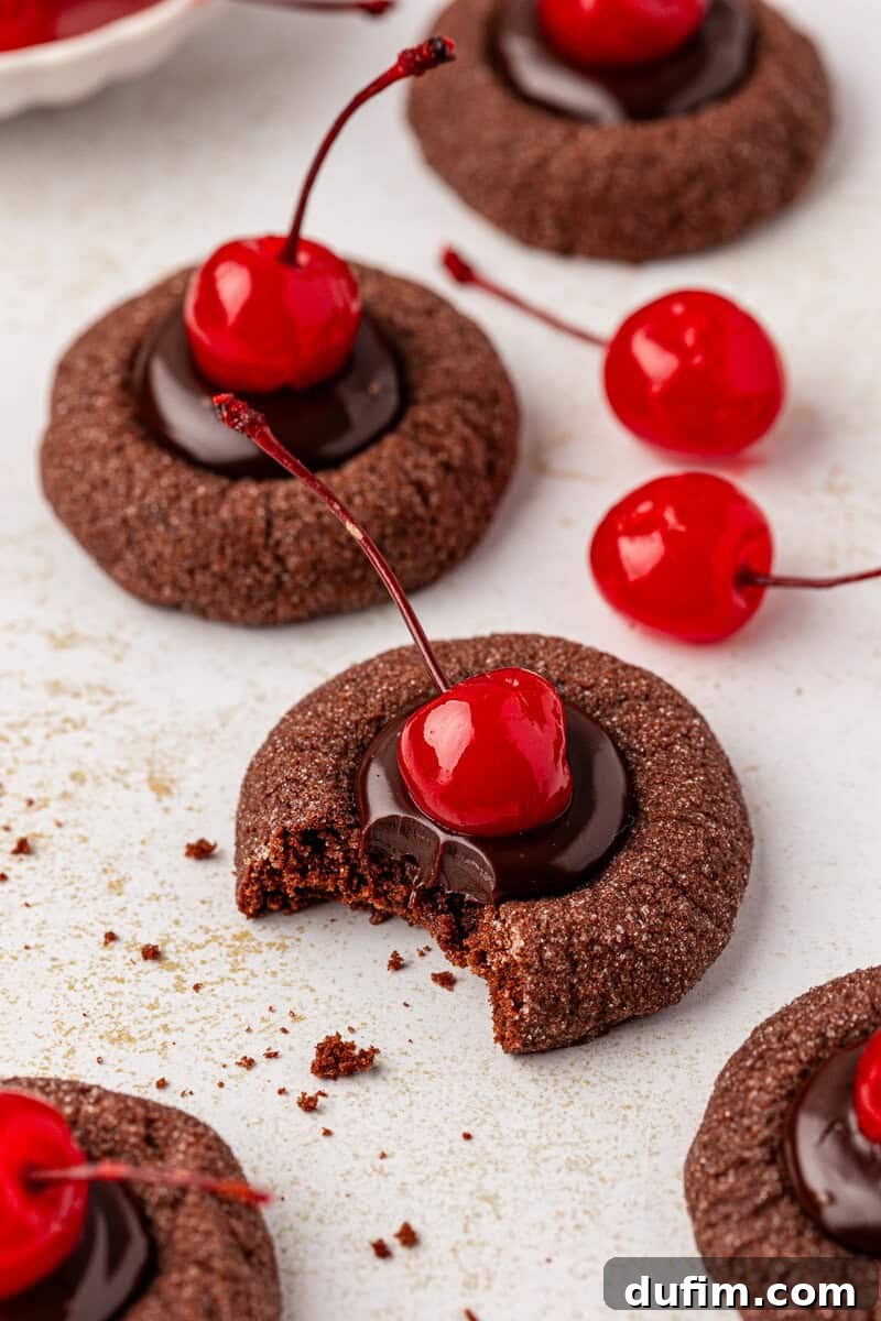 Close-up of freshly made Black Forest thumbprint cookies, each generously filled with chocolate ganache and crowned with a vibrant maraschino cherry. One cookie has a bite taken out, revealing its soft, dark chocolate texture. Other whole cookies and loose cherries are artfully arranged nearby on a white surface.