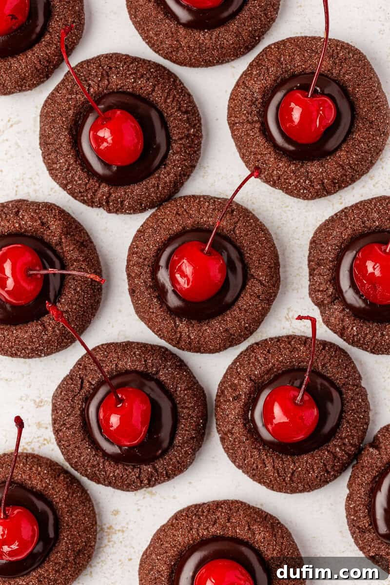 A close-up arrangement of delicious chocolate thumbprint cookies, each beautifully filled with glossy chocolate ganache and crowned with a bright red maraschino cherry. They are arranged closely together on a white surface, highlighting their rich color and inviting texture.