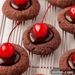 Several chocolate thumbprint cookies topped with glossy chocolate ganache and bright red maraschino cherries are arranged on a cooling rack.