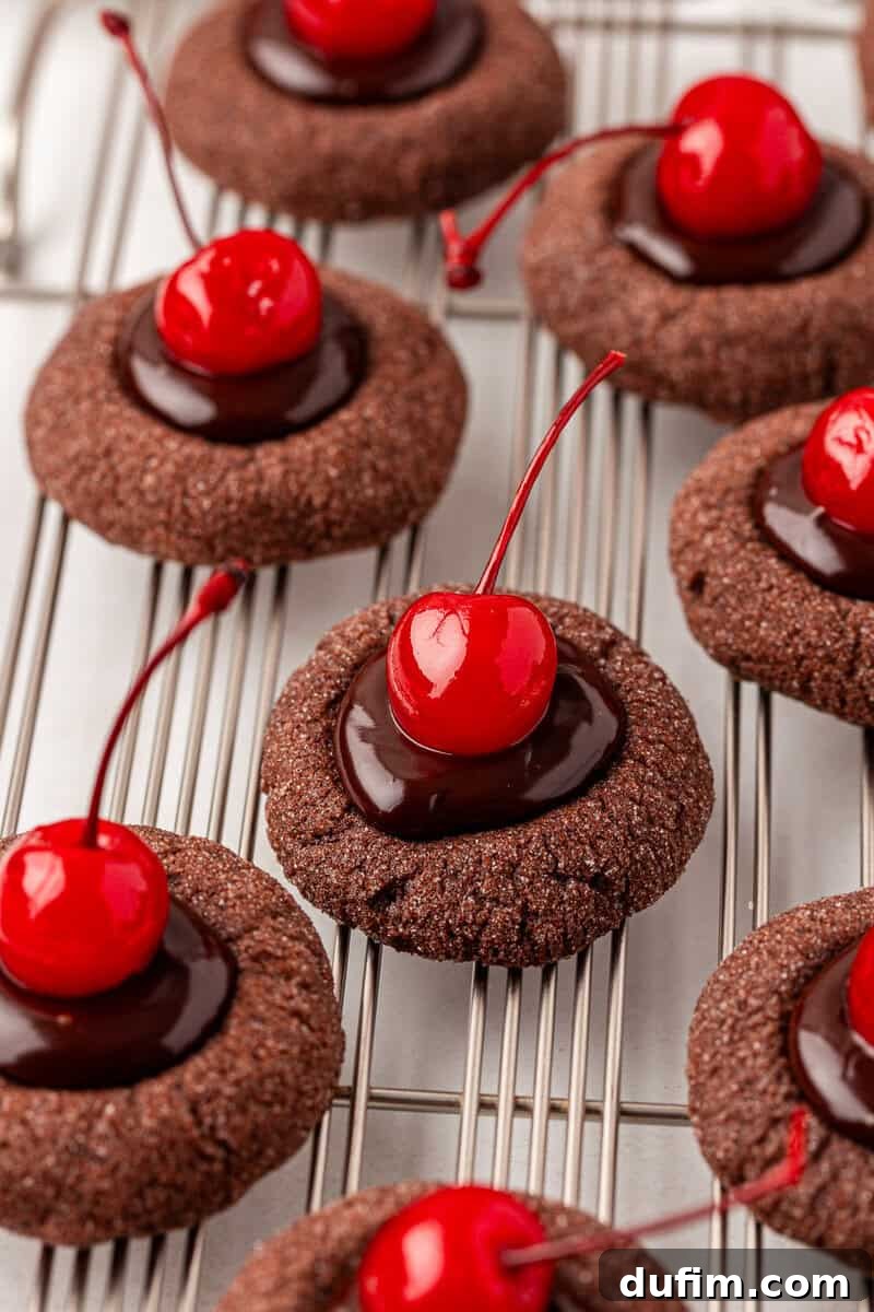Chocolate thumbprint cookies topped with glossy chocolate ganache and bright red maraschino cherries, arranged neatly on a metal cooling rack, showcasing their exquisite finish.