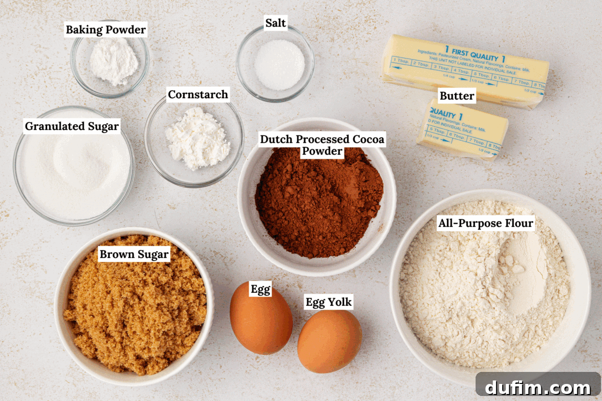 A top-down view of carefully labeled baking ingredients in individual bowls on a pristine white surface. Ingredients include baking powder, granulated sugar, brown sugar, cornstarch, salt, Dutch processed cocoa powder, all-purpose flour, two sticks of softened butter, a whole egg, and an isolated egg yolk.