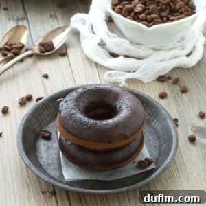 Two perfectly glazed coffee donuts, stacked on a metallic plate, surrounded by roasted coffee beans. A bowl of coffee beans and two spoons complete the rustic wooden table setting.