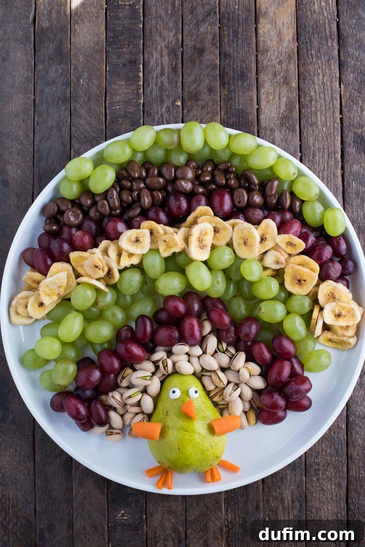 Vibrant turkey shaped fruit platter with pistachios, red and green grapes, banana chips, and a pear body with carrot details, all arranged on a large round white platter against a rustic wood background. The platter is full and colorful, showcasing the variety of fruits and nuts used for the turkey's tail feathers.
