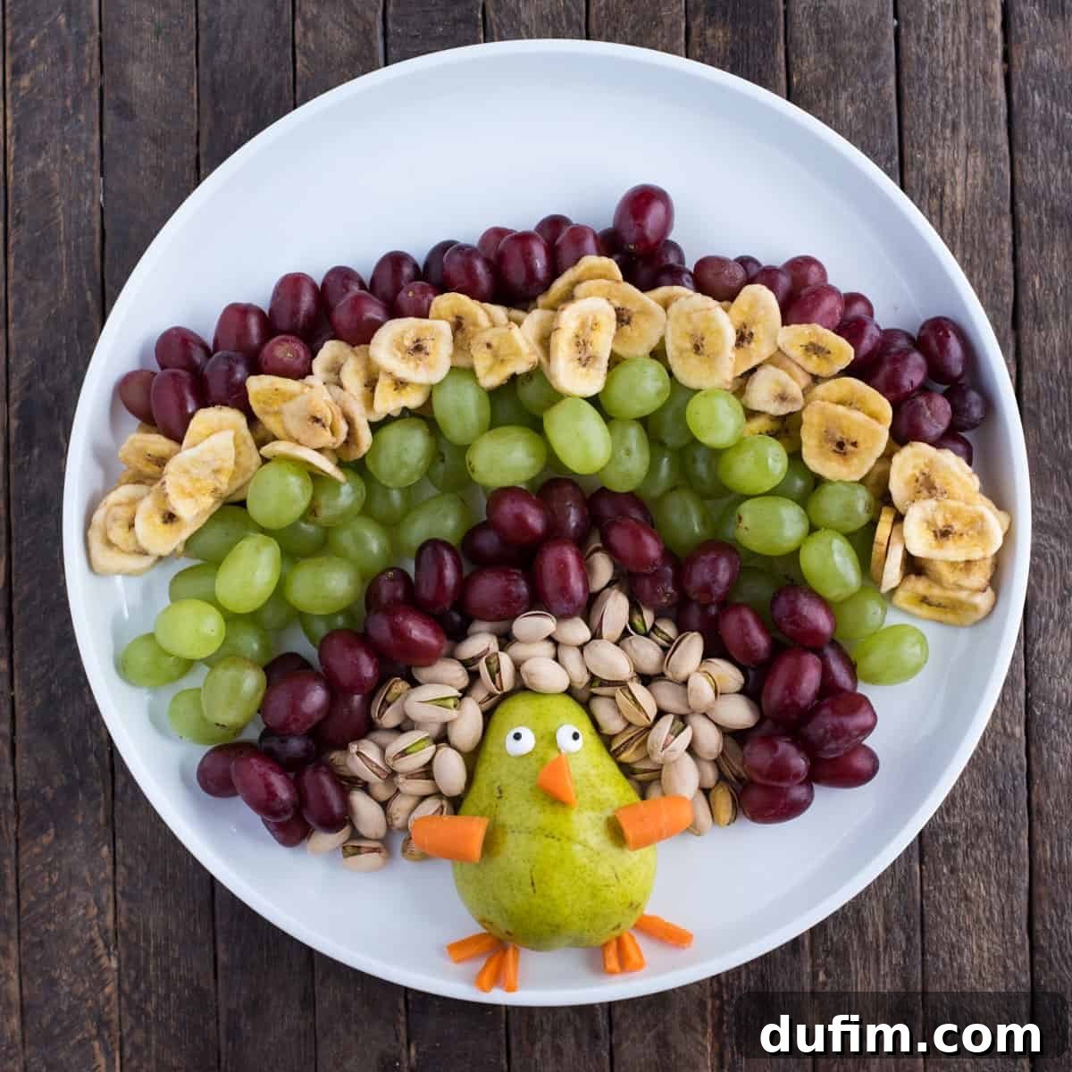 The turkey fruit platter progressing towards completion, featuring an additional layer of red grapes and the strategic placement of chocolate-covered raisins, which add both depth and a sweet element to the turkey's tail feathers. The layers are expanding outwards.