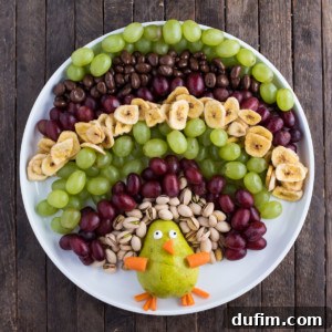 A beautifully arranged turkey fruit platter on a white plate, featuring a pear body, colorful grape feathers, pistachio accents, banana chips, and chocolate-covered raisins, set on a rustic wooden table. The platter is vibrant and inviting.