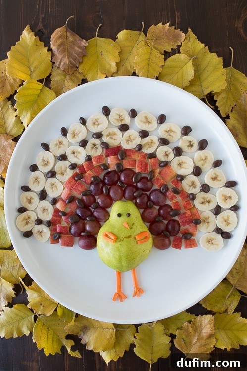 Close-up view of the turkey-shaped fruit platter, meticulously arranged on a pristine white serving tray. The pear body, playful candy eyes, carrot beak, and the vibrant layers of grape and nut 'feathers' are clearly visible, highlighting the artistic detail and freshness of the ingredients.