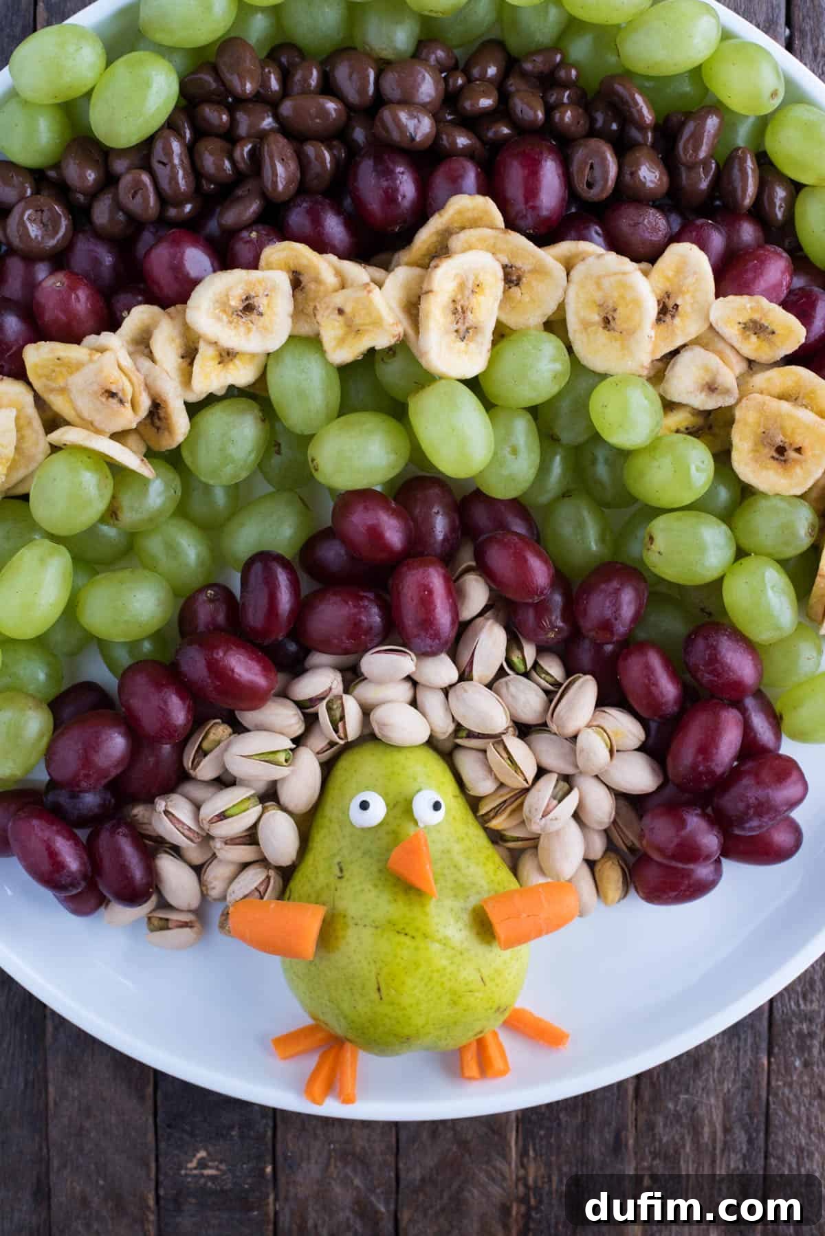 A flat lay photograph showcasing all the fresh and colorful ingredients required to assemble a turkey fruit platter. These include a halved pear, baby carrots, candy eyeballs, shelled pistachios, clusters of red and green grapes, banana chips, and chocolate-covered raisins, all arranged artfully on a large white round platter on a wooden surface, highlighting their readiness for assembly.