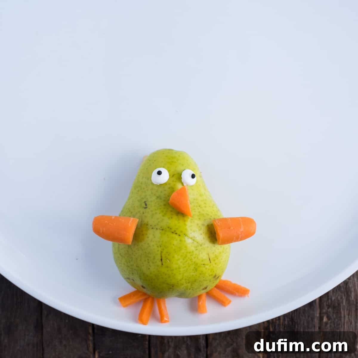 Step-by-step image showing a pear half placed on a platter with precisely carved carrot pieces for the turkey's beak, wings, and legs, illustrating the initial assembly phase before adding fruits. The pear is centered and the carrot pieces are clearly defined.