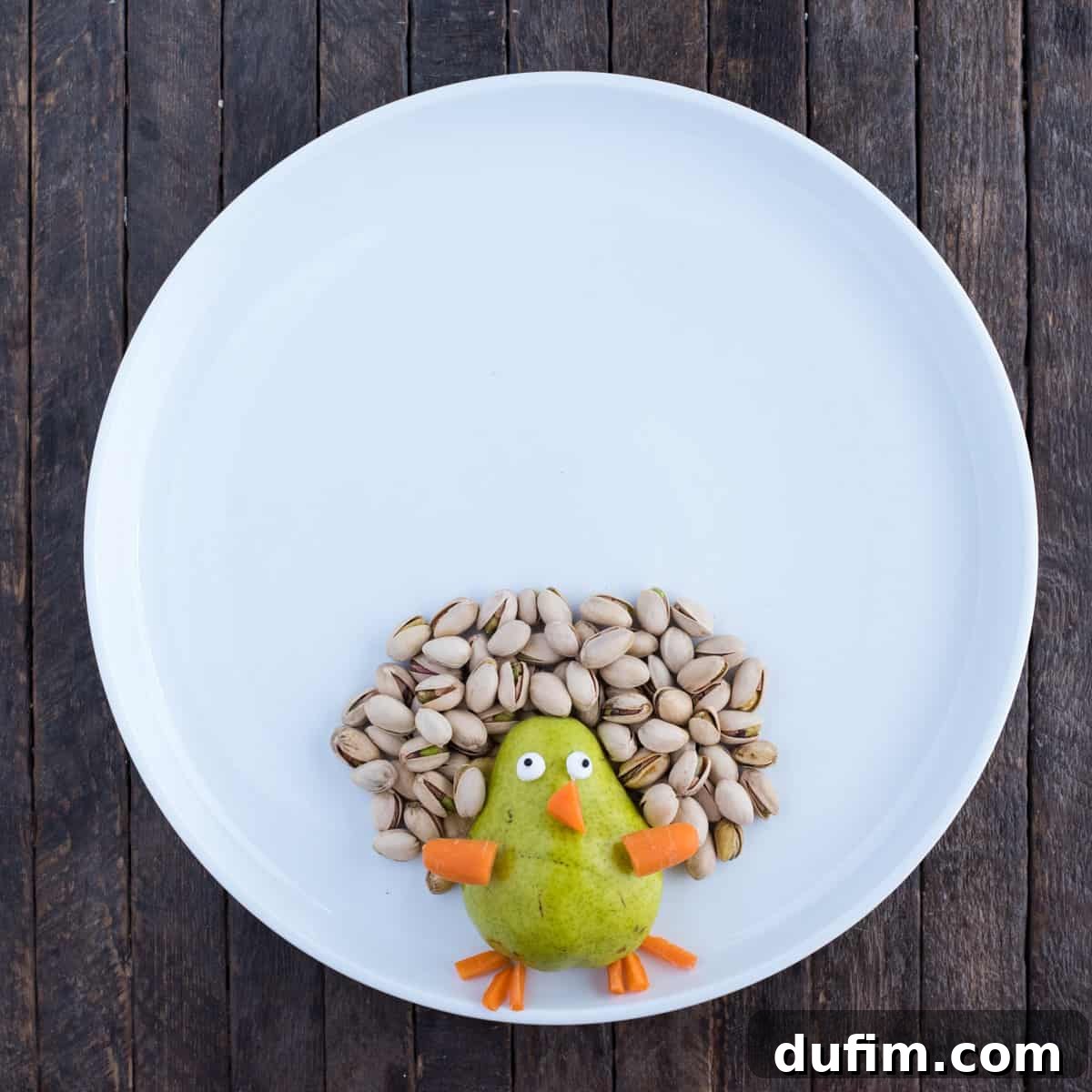 A step-by-step image of the fruit platter with the initial layers of turkey feathers. Shelled pistachios form an inner ring, followed by a vibrant outer ring of red grapes around the pear body, demonstrating the feathering process.