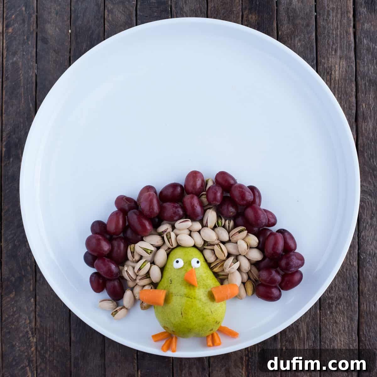 Another perspective of the fruit platter's progress, clearly showing the orderly arrangement of the first two layers of colorful feathers: bright green pistachios and juicy red grapes, creating a captivating halo around the pear turkey. The layers are tight and well-defined.