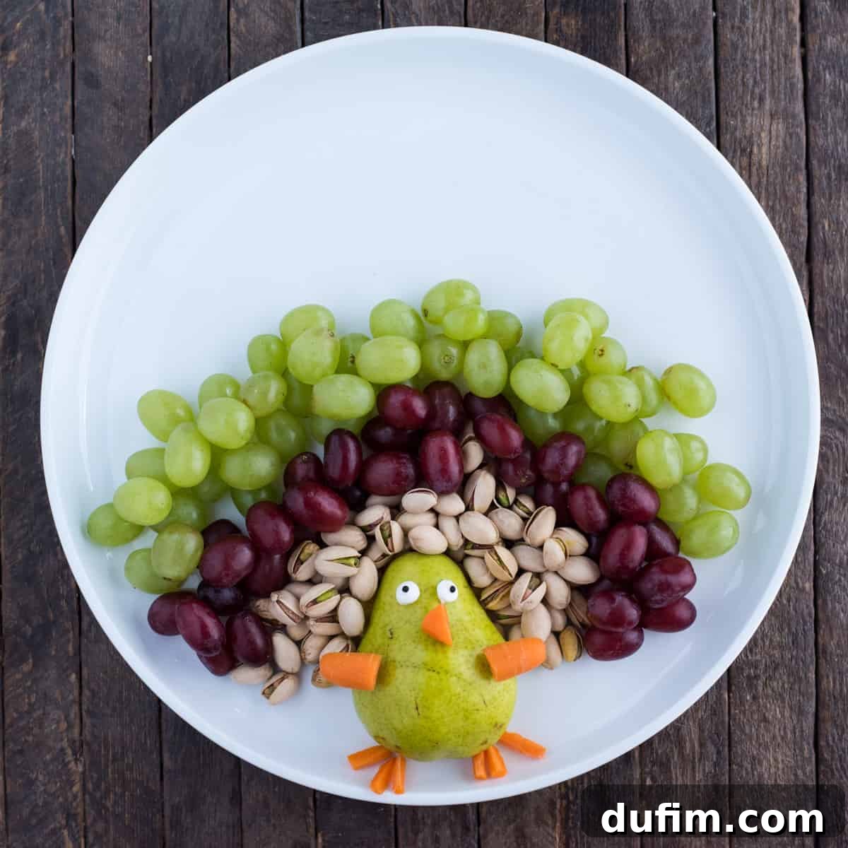 The turkey fruit platter demonstrating continued progress with the addition of green grapes and crunchy banana chips, further expanding the diverse and colorful feather pattern around the central pear turkey body. The arrangement shows clear distinct layers.