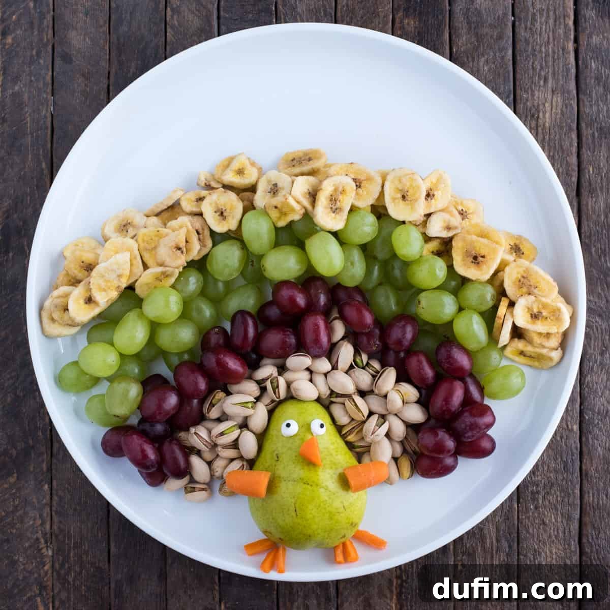A detailed close-up of the developing fruit turkey, clearly showcasing the distinct layers of red grapes, green grapes, and banana chips, highlighting the rich variety of textures and colors used in the feather arrangement. Each layer is neatly placed.