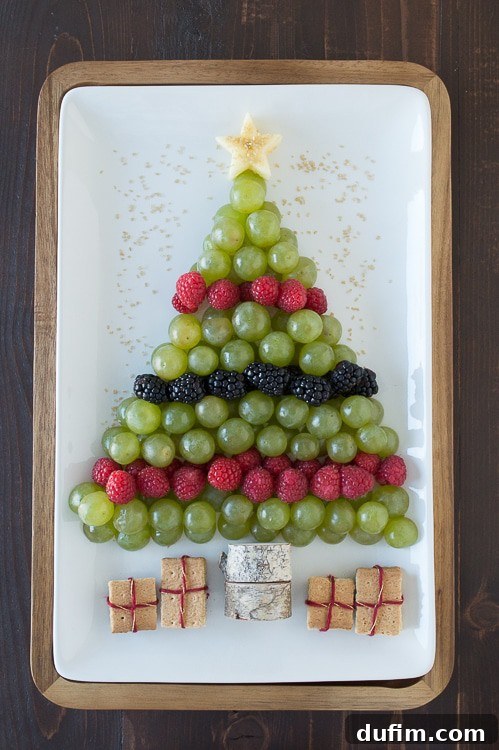 A vibrant Christmas tree fruit platter featuring green grapes, red raspberries, and dark blackberries, artfully arranged on a white platter. An apple star crowns the tree, with graham cracker presents nestled at the base.