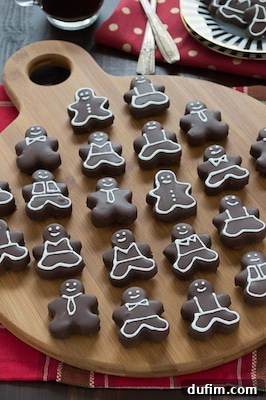 A wooden board displays rows of chocolate-covered gingerbread cookies shaped like people, each decorated with white icing to resemble various suits and smiling faces. In the background, a red napkin, utensils, and a slice of earthquake cake complete the scene.
