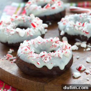 Chocolate donuts topped with white icing and crushed peppermint candies are arranged on a wooden board. Some candy cane donut pieces and peppermint bits are scattered around, creating a festive appearance.