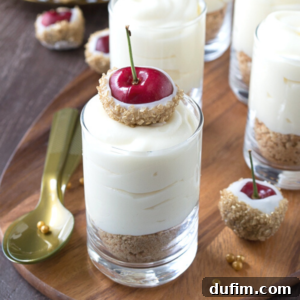 A white chocolate cheesecake shooter in a glass features a crumbly base, creamy white filling, and is topped with a cherry dipped in white chocolate and crushed nuts, displayed on a wooden tray with a gold spoon nearby.