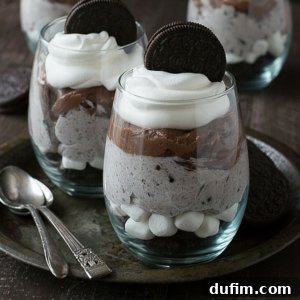 Two glass cups filled with layers of cookies and cream mousse, chocolate, whipped cream, and topped with a whole chocolate sandwich cookie—these Oreo parfaits rest on a metal tray with two spoons beside them.