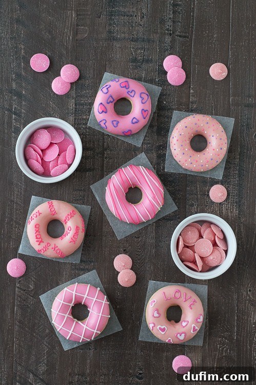 Six delicious Valentine’s Day Donuts alongside two white bowls containing different shades of pink candy melts, ready for decoration, on a wooden table.