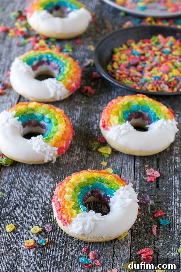 Four charming Rainbow Donuts, artfully decorated with rainbow-colored Fruity Pebbles, nestled amongst a scattering of colorful cereal on a warm wooden background.