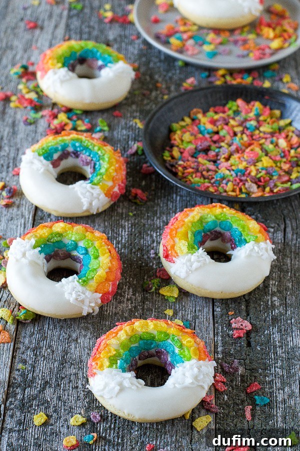 A close-up of five homemade Rainbow Donuts, artfully coated in white chocolate, adorned with shredded coconut clouds, and meticulously arranged Fruity Pebbles, all on a warm wooden surface.