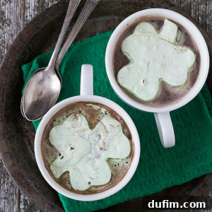Two mugs of hot chocolate topped with green shamrock marshmallows sit on a dark tray with two metal spoons and a green cloth napkin underneath, all on a rustic wooden surface.