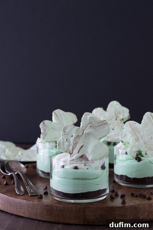 Shamrock Green Mint Chocolate Chip No-Bake Cheesecake 2 A beautifully layered No-Bake Mint Chocolate Chip Shamrock Cheesecake in a glass, with a dark cookie crumb base, vibrant green filling, and a topping of whipped cream and chocolate chips. Perfect for St. Patrick's Day.