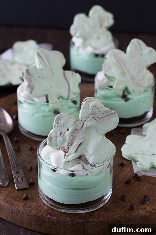 Shamrock Green Mint Chocolate Chip No-Bake Cheesecake 4 A trio of No-Bake Mint Chocolate Chip Shamrock Cheesecakes in individual glasses, beautifully arranged on a wooden board with extra chocolate chips scattered around. Each is topped with whipped cream and a sprinkle of mini chocolate chips, ready for a festive celebration.