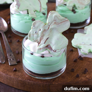 Shamrock Green Mint Chocolate Chip No-Bake Cheesecake 6 A glass dessert cup filled with layers of mint green mousse, reminiscent of shamrock cheesecake, topped with whipped cream, mini chocolate chips, and a shamrock-shaped meringue cookie. More desserts and chocolate chips are on a wooden tray in the background.