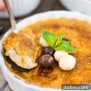 A close-up of espresso crème brûlée in a white ramekin, topped with a sprig of mint and three chocolate-coated candies. A spoon lifts a creamy portion, revealing the caramelized sugar crust.