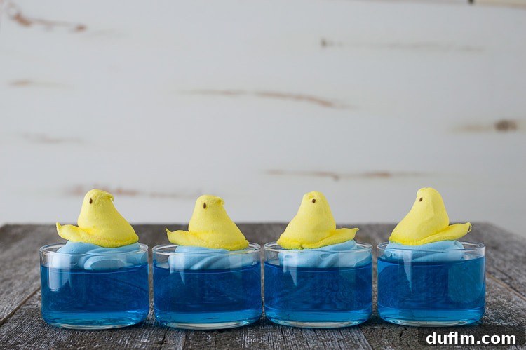 Four adorable Swimming Peeps Jello Cups arranged in a row, each featuring blue jello, blue whipped cream, and a yellow Peep chick, set against a rustic background.
