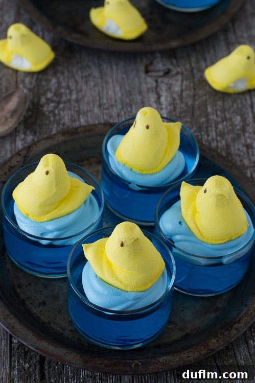 A single clear glass bowl filled with set blue jello, garnished with a swirl of light blue whipped cream and a bright yellow marshmallow Peep chick, ready for serving.