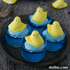 Four charming Peeps Jello Cups, featuring vibrant blue gelatin, swirls of light blue whipped cream, and yellow marshmallow chick candies, artfully arranged on a metal tray.