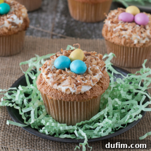 An angel food bird nest cupcake with toasted coconut, white frosting, and three pastel candy eggs sits on a black plate with green shredded paper, resembling a bird’s nest. Other similar cupcakes appear in the background.
