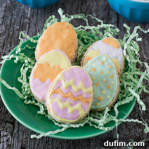 A green plate holds five Easter egg-shaped rice krispies treats decorated with pastel-colored icing in patterns like zigzags, dots, and stripes, placed on green shredded paper on a rustic wooden surface.