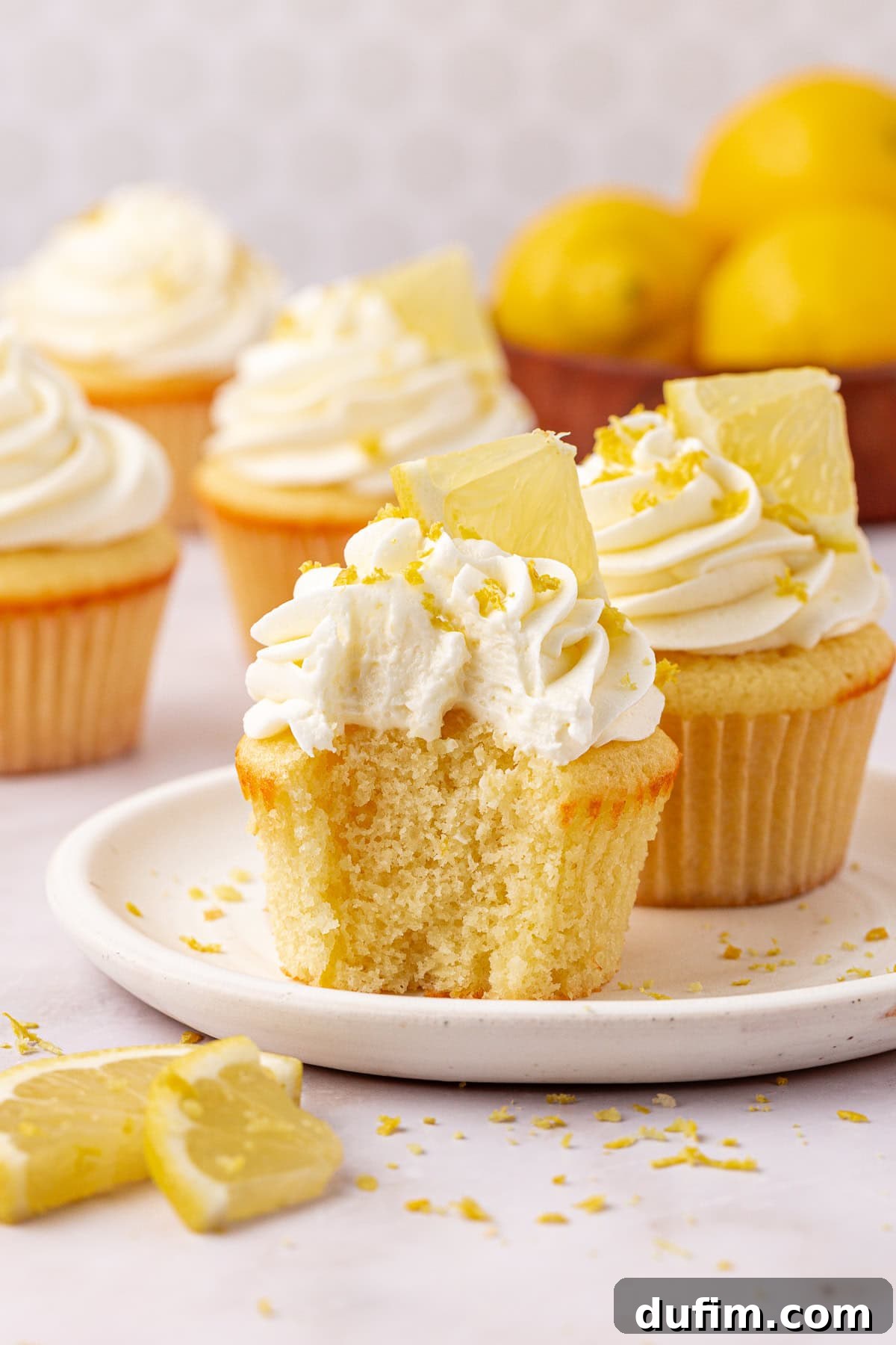 A plate with a lemon cupcake topped with whipped frosting and a lemon wedge; the cupcake has a bite taken off. More frosted cupcakes and a bowl of lemons are in the background. Lemon zest is sprinkled around.