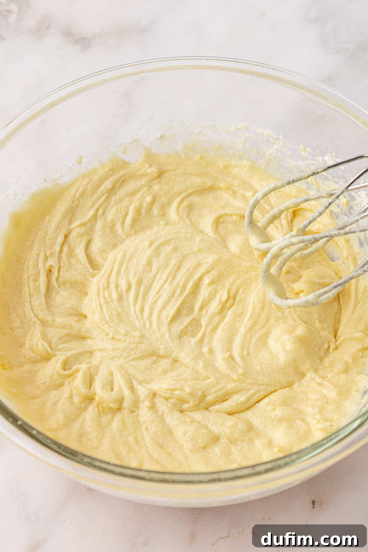A glass mixing bowl filled with creamy yellow cake batter sits on a marble surface. An electric hand mixer with batter on the beaters rests on the edge of the bowl.