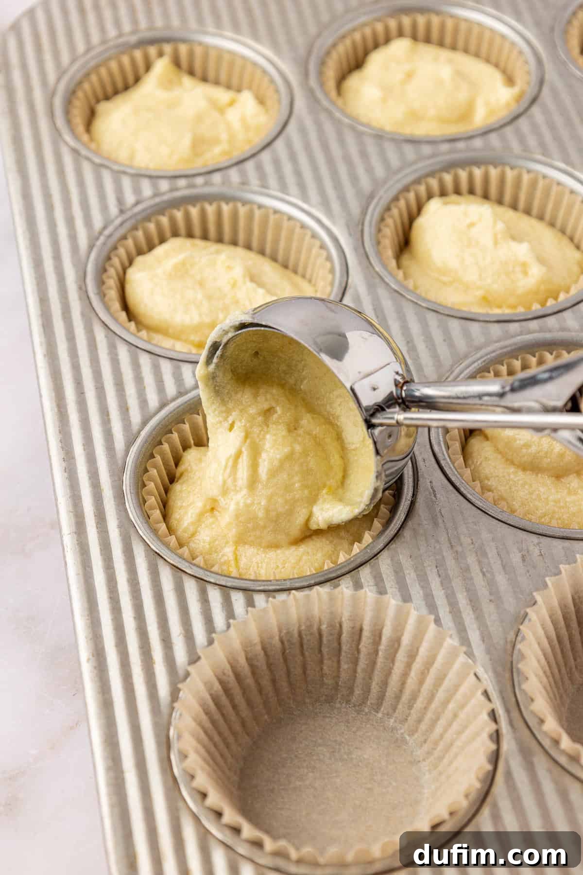 A metal ice cream scoop fills paper cupcake liners in a muffin tin with yellow batter; some liners are filled while others are still empty.