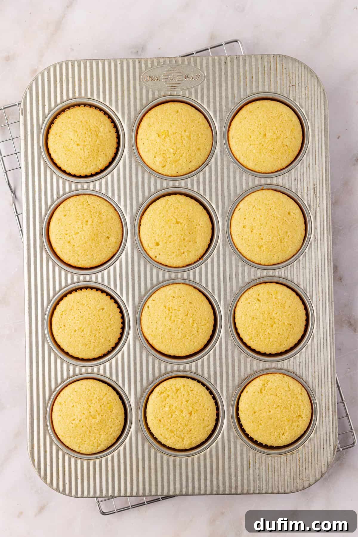 A metal muffin tin with twelve evenly baked yellow cupcakes in paper liners sits on a cooling rack atop a light-colored marble surface.