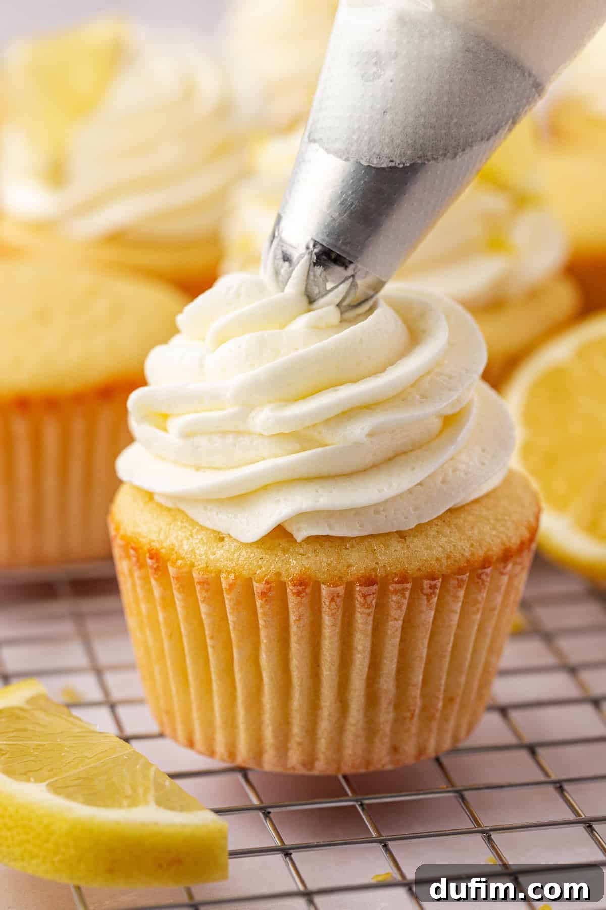 A close-up of a lemon cupcake on a cooling rack being topped with swirls of white frosting from a piping bag, with lemon slices in the background.