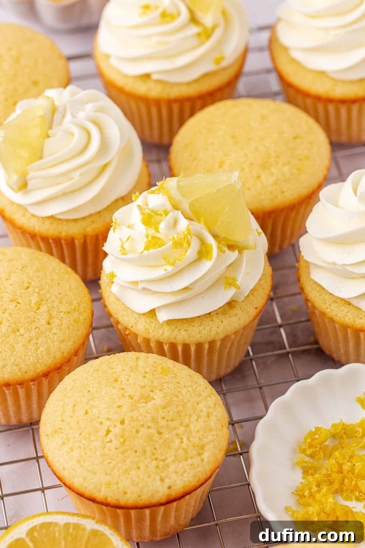Lemon cupcakes topped with swirls of white frosting, lemon zest, and lemon wedges are arranged on a cooling rack. Some cupcakes are unfrosted. A small dish of lemon zest is nearby.