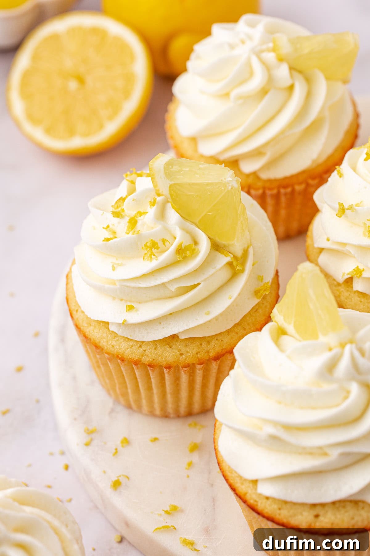 Four lemon cupcakes with swirled white frosting, topped with lemon slices and zest, are arranged on a white surface. In the background, there are halved lemons and additional lemon zest scattered around.