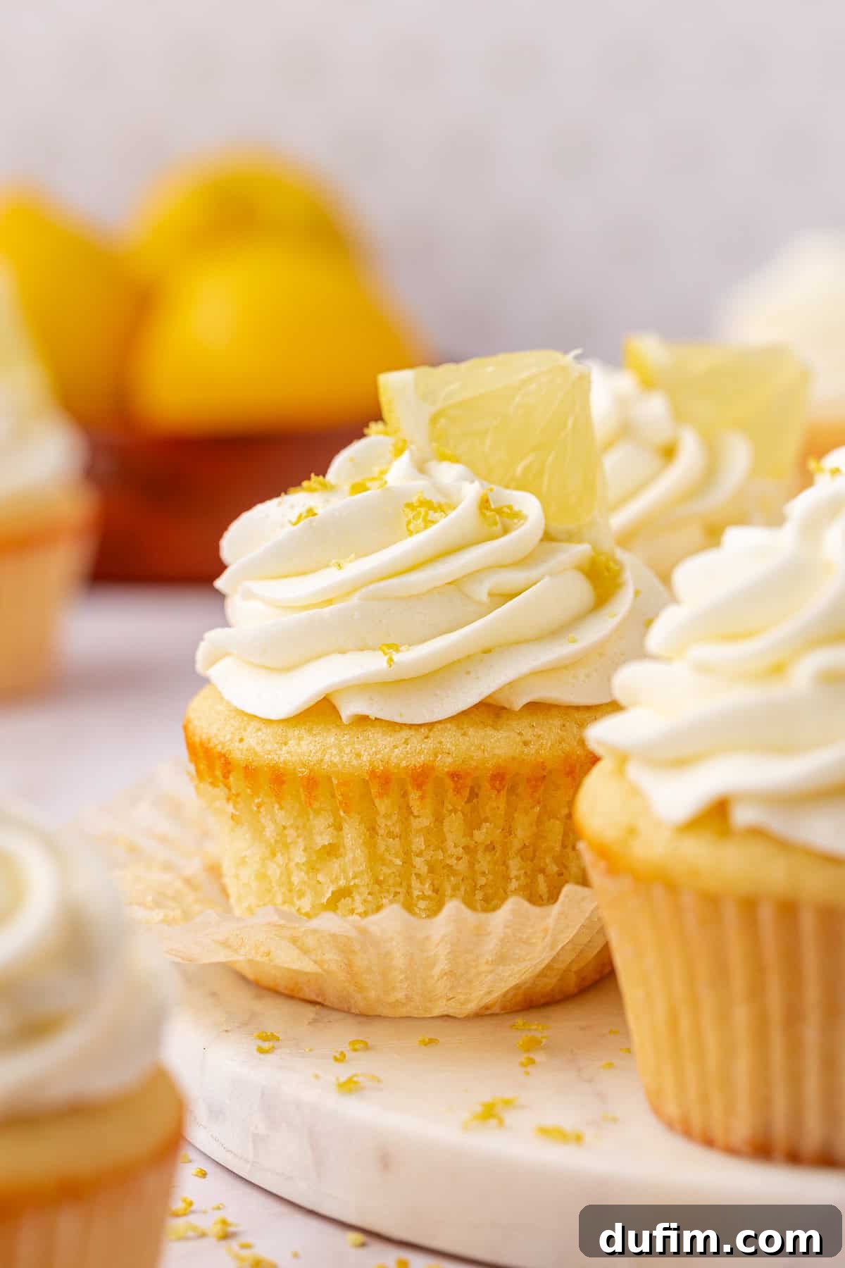 A close-up of lemon cupcakes with swirled white frosting, topped with lemon wedges and zest, sitting on a marble surface. In the background, whole lemons are slightly out of focus.
